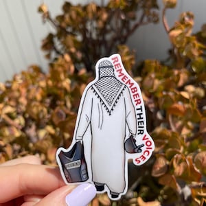 May include: A white sticker with a black and white illustration of a person wearing a traditional headscarf and a press vest. The person is holding a camera and a microphone. The sticker has the text "Remember Their Voices" written vertically on the right side.