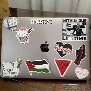 Peut inclure: Un ordinateur portable argent&eacute; avec divers autocollants, dont un drapeau palestinien, une colombe de la paix avec le texte "Free Palestine", un autocollant Hello Kitty et un autocollant avec le texte "From Palestine with love".