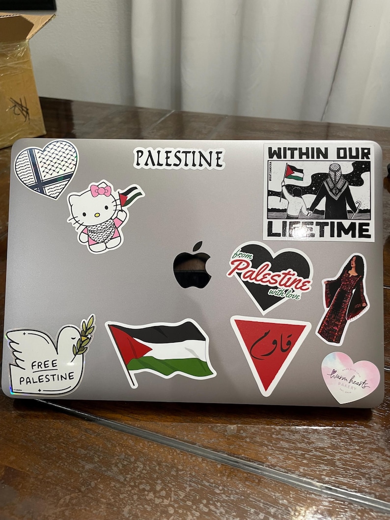 Peut inclure: Un ordinateur portable argent&eacute; avec divers autocollants, dont un drapeau palestinien, une colombe de la paix avec le texte "Free Palestine", un autocollant Hello Kitty et un autocollant avec le texte "From Palestine with love".