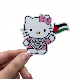 Peut inclure: Un personnage de Hello Kitty blanc portant une robe rose et un n&oelig;ud rose. Elle tient un drapeau palestinien dans sa main droite.