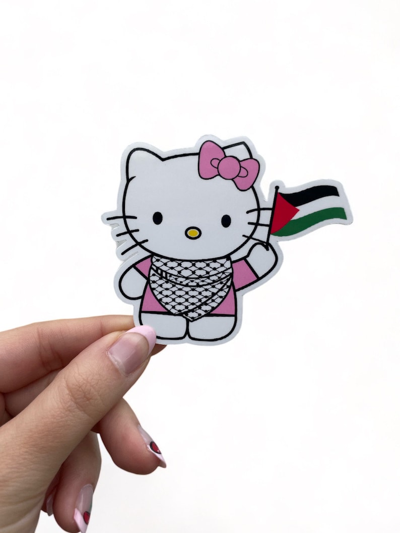 Peut inclure: Un personnage de Hello Kitty blanc portant une robe rose et un n&oelig;ud rose. Elle tient un drapeau palestinien dans sa main droite.