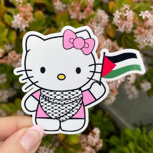 Peut inclure: Un personnage de Hello Kitty blanc portant une chemise rose et un keffieh noir et blanc. Elle tient un drapeau palestinien dans sa main droite. L'arri&egrave;re-plan est une image floue de plantes vertes avec des fleurs blanches.