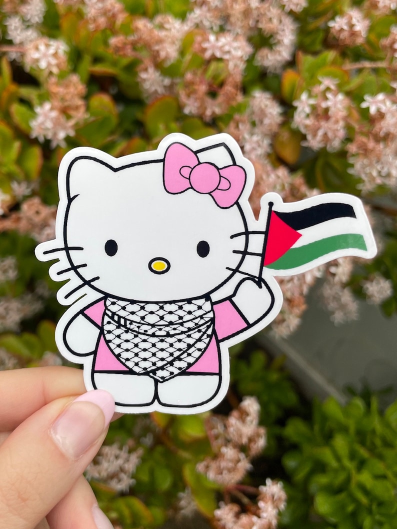 Peut inclure: Un personnage de Hello Kitty blanc portant une chemise rose et un keffieh noir et blanc. Elle tient un drapeau palestinien dans sa main droite. L'arri&egrave;re-plan est une image floue de plantes vertes avec des fleurs blanches.