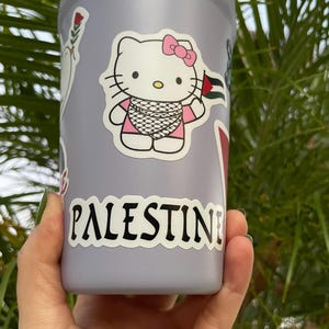 Peut inclure: Une bouteille d'eau violette avec un autocollant Hello Kitty portant un keffieh et tenant un drapeau palestinien. La bouteille a &eacute;galement un autocollant qui dit "PALESTINE" en lettres noires.