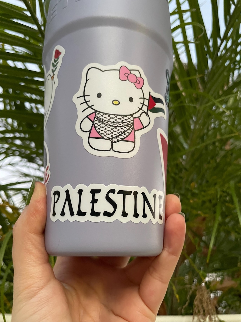Peut inclure: Une bouteille d'eau violette avec un autocollant Hello Kitty portant un keffieh et tenant un drapeau palestinien. La bouteille a &eacute;galement un autocollant qui dit "PALESTINE" en lettres noires.