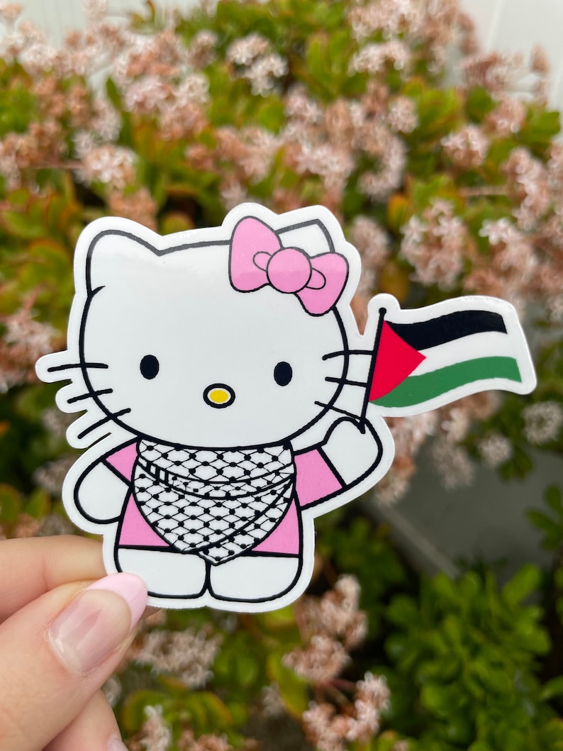 Peut inclure: Un personnage de dessin anim&eacute; Hello Kitty blanc portant une robe rose et un keffieh noir et blanc. Le personnage tient un drapeau palestinien.