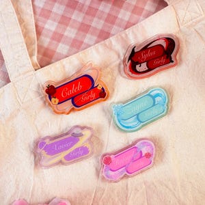 Op de afbeelding: Een verzameling heldere acryl naamlabels met de namen Caleb, Sylus, Zayne, Xavier en Rafayel. Elke badge heeft een uniek ontwerp en het woord "Girly". De badges liggen op een canvas draagtas.