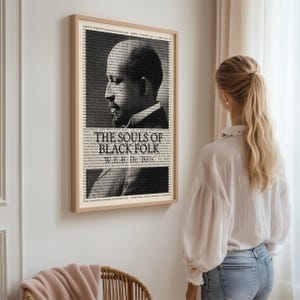 May include: A framed black and white print of W.E.B. Du Bois, with the title "The Souls of Black Folk." The portrait is set against a backdrop of text, with a light brown wooden frame. The print is hanging on a white wall.