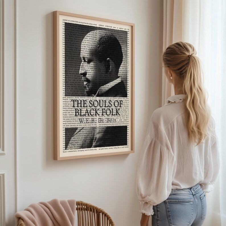 May include: A framed black and white print of W.E.B. Du Bois, with the title "The Souls of Black Folk." The portrait is set against a backdrop of text, with a light brown wooden frame. The print is hanging on a white wall.