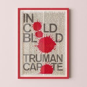 May include: A framed print with a red border displays the text "IN COLD BLOOD TRUMAN CAPOTE" in large, dark letters over a background of printed text. Red paint splatters are overlaid on the text.