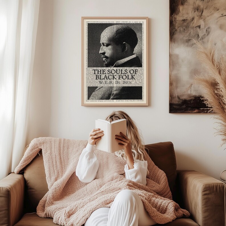 May include: A framed black and white print of W.E.B. Du Bois with the text "The Souls of Black Folk" hangs on a wall. A person is seated on a brown sofa, reading a book and covered with a pink blanket.