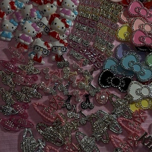 May include: A collection of assorted decorative charms and hair accessories featuring Hello Kitty figures, rhinestone-encrusted planet shapes, colorful bows, floral motifs, and small cherry designs, all arranged together on a textured pink surface.