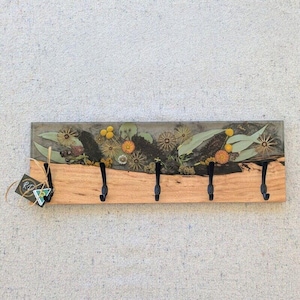 May include: A wooden key holder with five black hooks. The top section is a clear resin with embedded dried flowers and leaves. The bottom section is natural wood. A tag hangs from the left side.