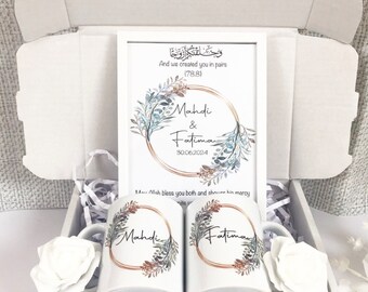 May include: A white gift box with a white frame inside. The frame has a watercolor floral design with the names Mahdi & Fatima and the date 30.06.2024. The box also contains two white mugs with the same floral design and the names Mahdi and Fatima. The text in the frame reads "And we created you in pairs (78.8)" and "May Allah bless you both and shower his mercy".