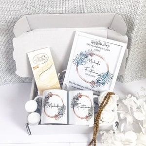 Könnte beinhalten: Eine weiße Geschenkbox gefüllt mit verschiedenen Artikeln, darunter zwei weiße Tassen mit goldenen Verzierungen und Blumendesigns, ein gerahmter Druck mit einem Blumendesign und dem Text "Mahdi & Fatima 30.06.2024", eine Schokoladentafel und eine goldene Perlenkette.