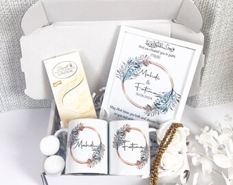 May include: A white gift box filled with a variety of items, including two white mugs with gold trim and floral designs, a framed print with a floral design and the text "Mahdi & Fatima 30.06.2024", a chocolate bar, and a gold beaded necklace.