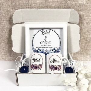 Könnte beinhalten: Eine weiße Geschenkbox mit zwei Tassen, einem gerahmten Druck und zwei Kerzen. Die Tassen sind mit Blumendesigns und den Namen "Bilal" und "Alina" verziert. Der gerahmte Druck zeigt die Namen "Bilal & Alina" und das Datum "2. März 2024".