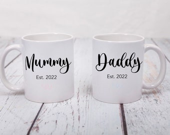 May include: Two white ceramic mugs with black lettering. The mug on the left says "Mummy Est. 2022" and the mug on the right says "Daddy Est. 2022".