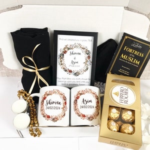 Könnte beinhalten: Eine weiße Geschenkbox gefüllt mit verschiedenen Artikeln, darunter zwei weiße Tassen mit den Namen "Shirina" und "Azim" und dem Datum "24/02/2024" darauf gedruckt, ein schwarzer Schal, ein gerahmter Druck mit einem Blumenkranz und dem Text "And we created you in pairs 78.8", ein Buch mit dem Titel "Fortress of the Muslim" und eine Schachtel Ferrero Rocher Pralinen.