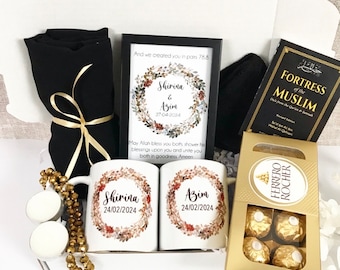 May include: A white gift box filled with a variety of items, including two white mugs with the names "Shirina" and "Azim" and the date "24/02/2024" printed on them, a black scarf, a framed print with a floral wreath and the text "And we created you in pairs 78.8", a book titled "Fortress of the Muslim", and a box of Ferrero Rocher chocolates.