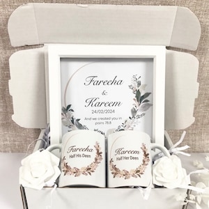 Könnte beinhalten: Eine weiße Geschenkbox mit einem gerahmten Druck und zwei Tassen. Der Druck lautet "Fareeha & Kareem 24/02/2024" mit floralen Akzenten. Auf den Tassen steht "Fareeha Half His Deen" und "Kareem Half Her Deen". Weiße Rosen sind ebenfalls in der Box.