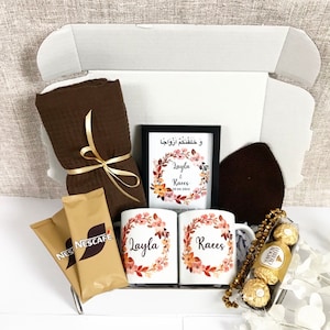 Könnte beinhalten: Eine Geschenkbox gefüllt mit Artikeln für eine Hochzeitsfeier. Die Box enthält zwei Tassen mit den Namen "Layla" und "Raees" darauf, eine braune Decke, zwei Päckchen Instantkaffee, einen gerahmten Druck mit den Namen "Layla & Raees" und dem Datum "20.04.2024", eine braune Strickmütze, eine goldene Perlenkette und eine Schachtel Pralinen.