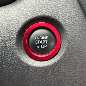 Puede incluir: Primer plano del botón de arranque/parada del motor de un coche. El botón negro tiene el texto blanco "ENGINE START STOP". Está rodeado por un anillo rojo brillante, encastrado en un salpicadero gris oscuro.
