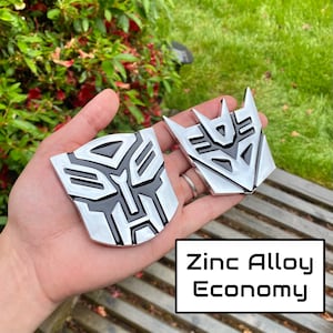May include: Two silver and black zinc alloy emblems with the Transformers logo. The emblems are held in a hand, with the text "Zinc Alloy Economy" in a white box.