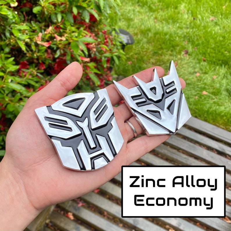 May include: Two silver and black zinc alloy emblems with the Transformers logo. The emblems are held in a hand, with the text "Zinc Alloy Economy" in a white box.