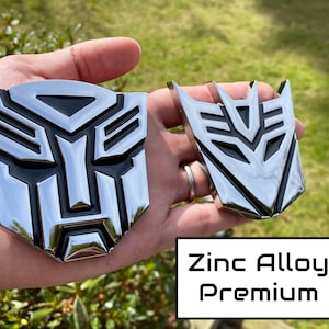 May include: Two chrome-finished zinc alloy emblems, one larger and one smaller, featuring a stylized geometric design with black accents. The emblems are displayed against a natural green background. The text "Zinc Alloy Premium" is visible.