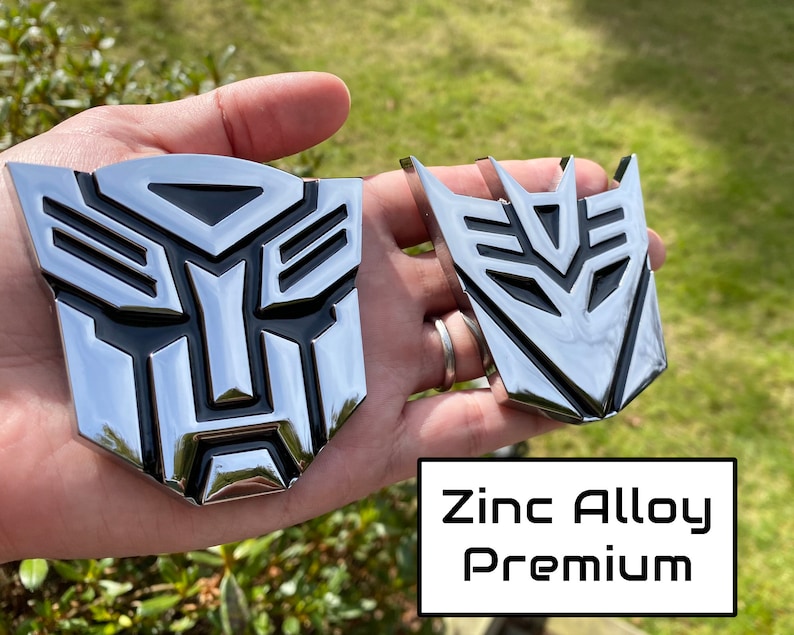 May include: Two chrome-finished zinc alloy emblems, one larger and one smaller, featuring a stylized geometric design with black accents. The emblems are displayed against a natural green background. The text "Zinc Alloy Premium" is visible.