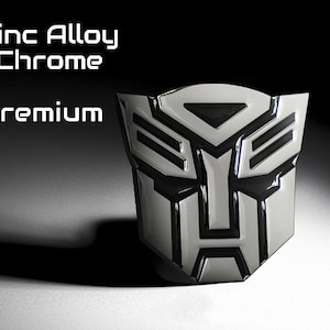 May include: A chrome-finished emblem with a black and silver design. The emblem features a stylized face with geometric shapes. The text "Zinc Alloy Chrome Premium" is displayed above the emblem. The emblem is likely for automotive or collectible purposes.