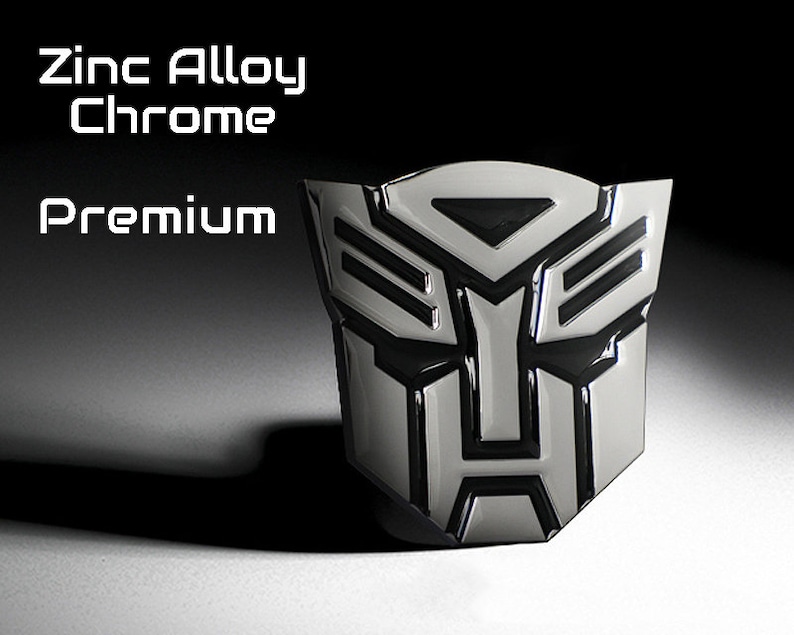 May include: A chrome-finished emblem with a black and silver design. The emblem features a stylized face with geometric shapes. The text "Zinc Alloy Chrome Premium" is displayed above the emblem. The emblem is likely for automotive or collectible purposes.