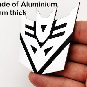 May include: A white and black emblem made of aluminum, 2mm thick. The emblem has a geometric design with sharp angles and is held in a hand. The text "Made of Aluminium" and "2mm thick" is visible.