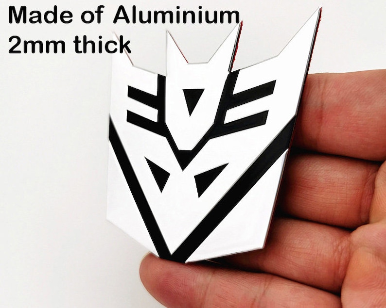May include: A white and black emblem made of aluminum, 2mm thick. The emblem has a geometric design with sharp angles and is held in a hand. The text "Made of Aluminium" and "2mm thick" is visible.