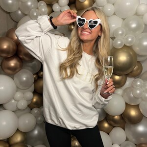 Pode incluir: Uma mulher vestindo uma camiseta branca com a palavra "Bride" nela, leggings pretos e &oacute;culos de sol em forma de cora&ccedil;&atilde;o que dizem "2023 The Wedding" em letras pretas. Ela est&aacute; segurando uma ta&ccedil;a de champanhe e sorrindo. O fundo &eacute; uma parede de bal&otilde;es brancos e dourados.