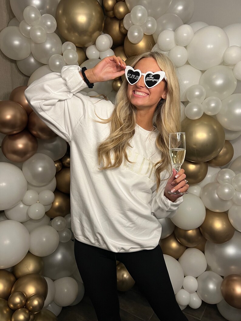 Pode incluir: Uma mulher vestindo uma camiseta branca com a palavra "Bride" nela, leggings pretos e &oacute;culos de sol em forma de cora&ccedil;&atilde;o que dizem "2023 The Wedding" em letras pretas. Ela est&aacute; segurando uma ta&ccedil;a de champanhe e sorrindo. O fundo &eacute; uma parede de bal&otilde;es brancos e dourados.