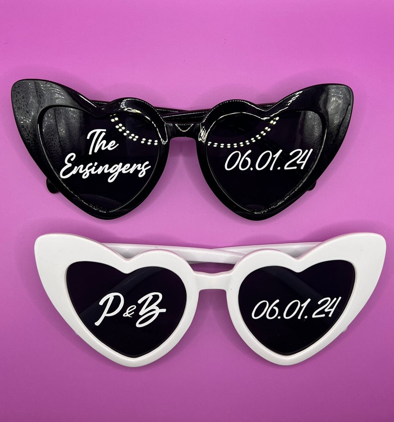 Pode incluir: Dois pares de &oacute;culos de sol em forma de cora&ccedil;&atilde;o, um preto e um branco. O preto tem o texto "The Ensingers" e "06.01.24" nas lentes. O branco tem o texto "P&B" e "06.01.24" nas lentes.