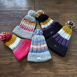 May include: Five colourful knitted winter hats with pom-poms. The hats are striped with various colours including red, pink, yellow, white, blue, purple, and black.