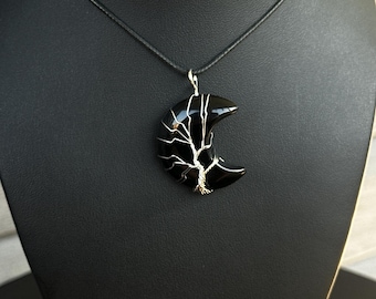 May include: A black crescent moon pendant made of polished stone, wrapped in silver-coloured wire shaped like a tree with branches extending across the surface. The pendant hangs from a simple black cord necklace, creating a contrast between the dark stone and the metallic wire design.