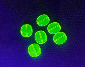 May include: Six luminous, oval-shaped, neon green beads with a single groove across the centre, arranged on a dark blue fabric. The beads are evenly spaced and appear translucent.