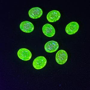 May include: Ten oval neon green glass beads, each with a floral design at the centre. The beads are scattered on a black surface, creating a striking contrast. The beads are likely for jewellery making.