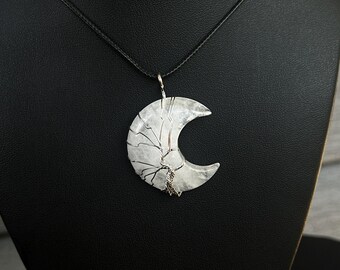 May include: A crescent moon-shaped pendant crafted from a translucent white stone, featuring a silver-toned wire-wrapped tree design across its surface. The pendant hangs from a simple black cord, creating a clear contrast against the light stone.