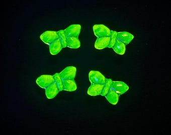 May include: Four small butterfly-shaped objects with a vibrant, glowing neon green finish. Each butterfly features symmetrical wings and a central body detail, presented against a solid black background, creating a high-contrast visual effect.