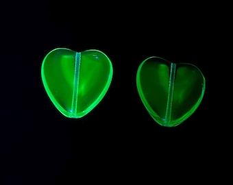 May include: Two heart-shaped beads that glow with a bright neon green light against a solid black background. Each bead features a vertical channel running through the centre, creating a distinct line that divides the heart shape into two halves.