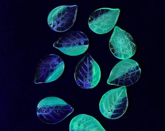 May include: Ten leaf-shaped glass beads in a teal hue with internal purple veining. The beads are arranged on a dark blue background. These translucent beads are likely for crafting jewellery or other decorative items. The beads are a striking teal colour.
