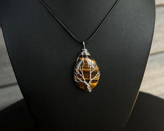 May include: A teardrop-shaped tiger's eye gemstone pendant wrapped in silver-coloured wire shaped like a tree. The pendant hangs from a simple black cord necklace. The stone features horizontal bands of golden-brown and dark brown colours.