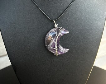 May include: A crescent moon-shaped pendant crafted from purple and white banded stone, wrapped in silver-coloured wire arranged in a tree-like pattern. The pendant hangs from a simple black cord necklace, showcasing the natural colour variations.