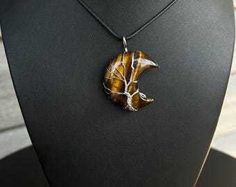 May include: A crescent moon-shaped tiger's eye pendant wrapped in silver-coloured wire to resemble a tree, suspended from a simple black cord necklace. The stone features characteristic golden-brown bands and a polished, reflective surface finish.