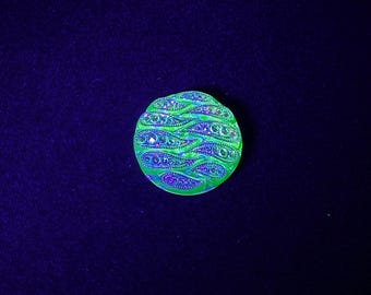 May include: A circular decorative object in green and purple, featuring a paisley pattern. The object has a textured surface and is set against a dark blue background. The object is round in shape.